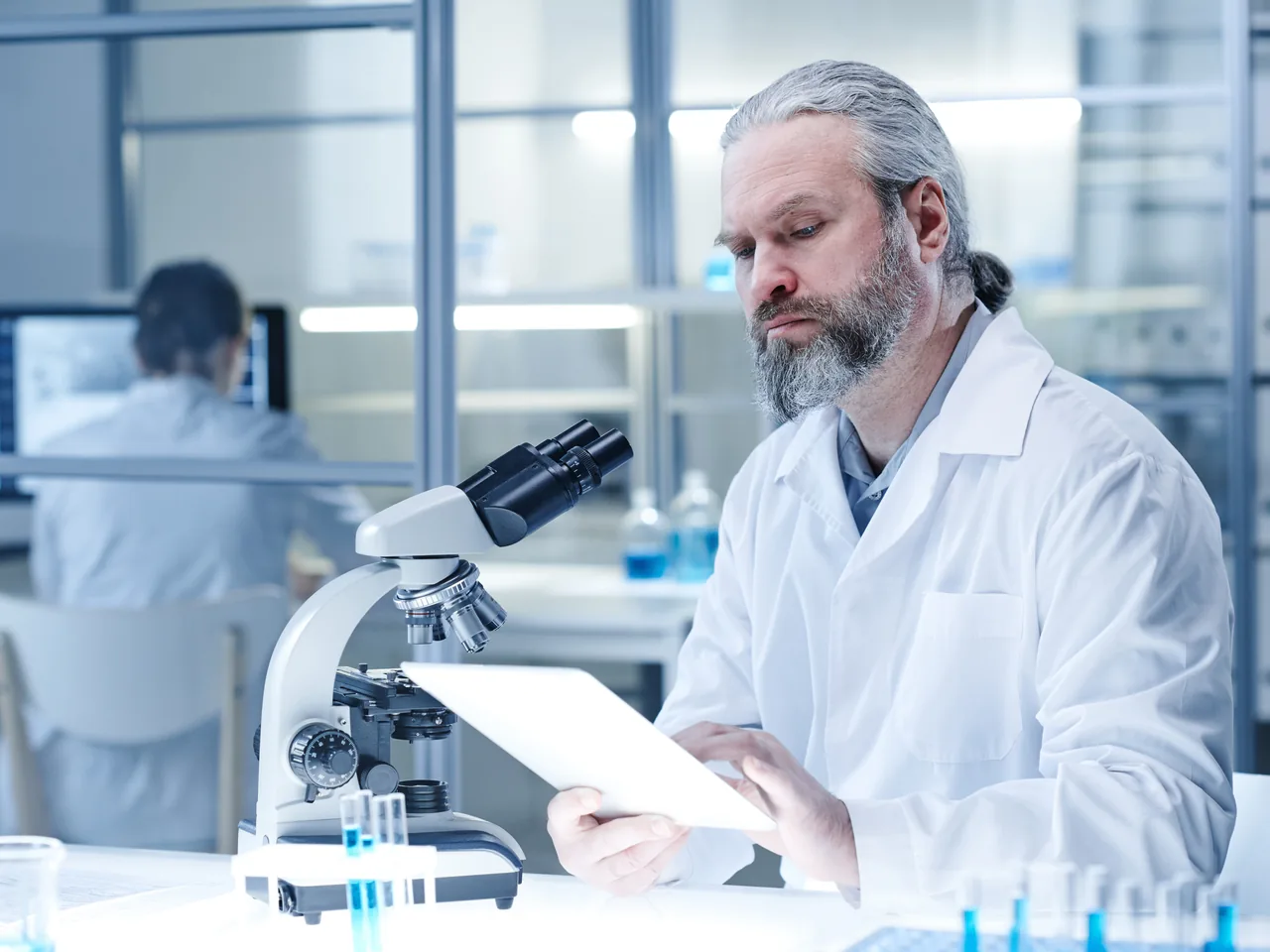 Scientist in a white lab coat working on a tablet next to a microscope
